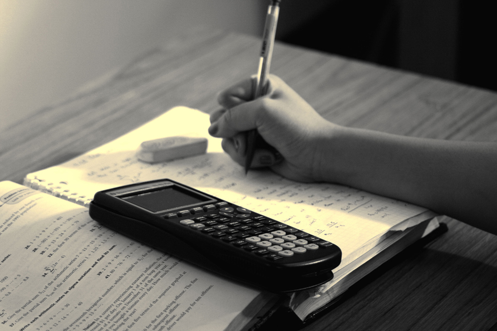 Close-up photo of a hand writing in a notebook. On top of the notebook is a graphing calculator.