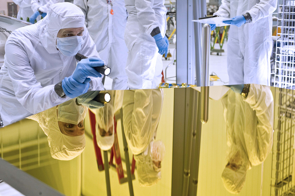 People in white clothing covered from head to toe and wearing blue colored gloves are working in a research laboratory setting, one person holding a flash light and analyzing and another reading a manuscript and so on. Their images are seen on a smooth colored glass top of a work table.