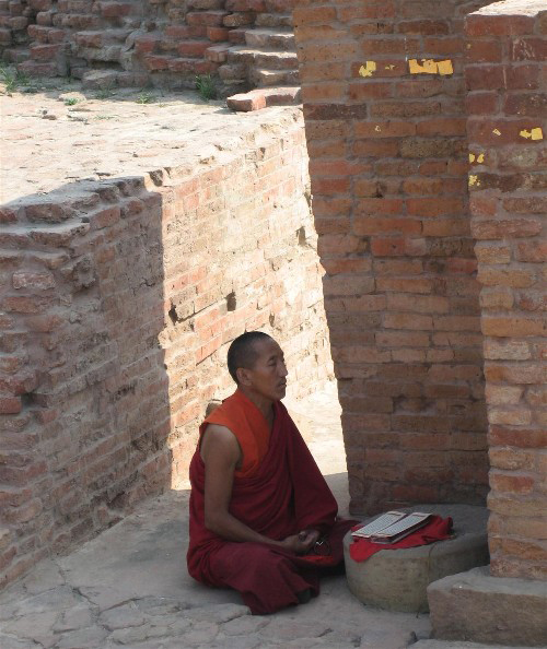 A man dressed in an orange robe is shown with his legs crosses, sitting within outdoor brick walls.