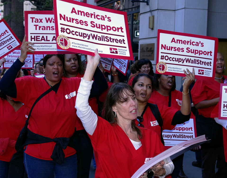 Several women dressed in red nurses’ scrubs march and hold signs reading America’s Nurses Support at Occupy Wall Street above their heads.