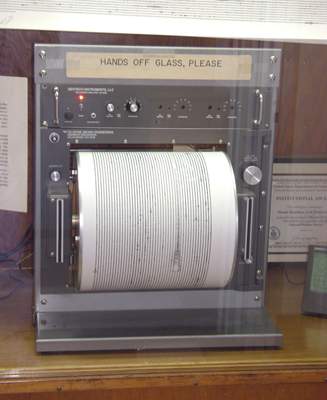 The figure shows a seismograph put on a wooden table. Its top is labeled as “Hands off glass, please”. Below it there are some buttons are shown and a paper roller is fitted in the seismograph to print the observation by the machine. On the right and left of the roller, two vertical cable slots are given.