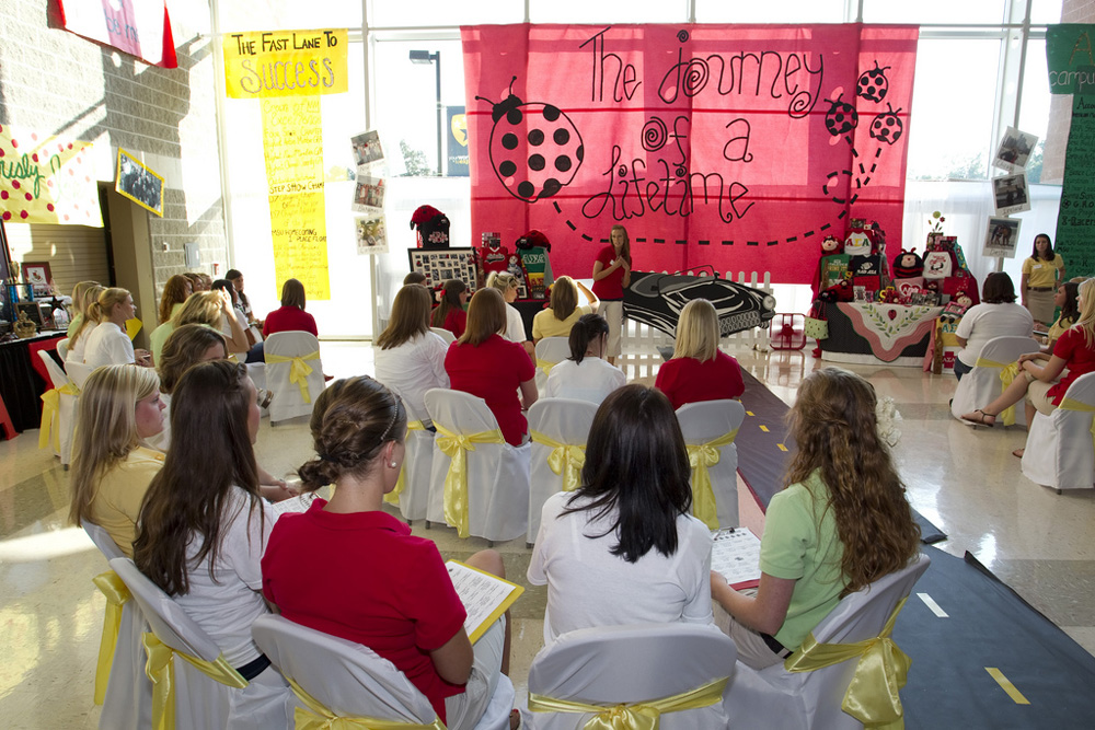 About a dozen young females are shown sitting in chairs at a sorority recruitment on campus.