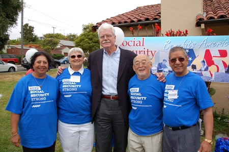 A tall man with white hair and moustache and glasses in casual business attire is shown flanked by two elderly women on his right and two elderly men on his left. The elderly people are all wearing blue T-shirts reading “Keep Social Security Strong: A A R P.” A banner in the background can also be seen, reading “Social Security Benefits America.”