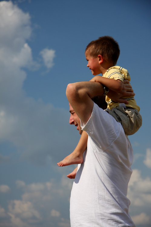 A man and boy are shown riding bicycles along a path in a park, man with hand on boy's shoulder.