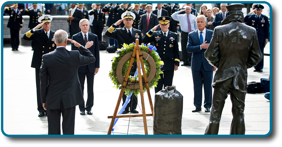 A photograph shows several key members of the United States military accompanied by a crowd as they stand facing toward a wreath. All hold their right arms in salute or placed across their chests.