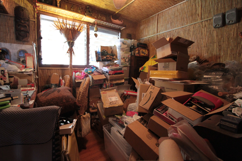 A photograph shows a small room containing tall piles of boxes, overflowing with papers, binders, and various other possessions. Much of the furniture and floor are concealed beneath these other objects. 