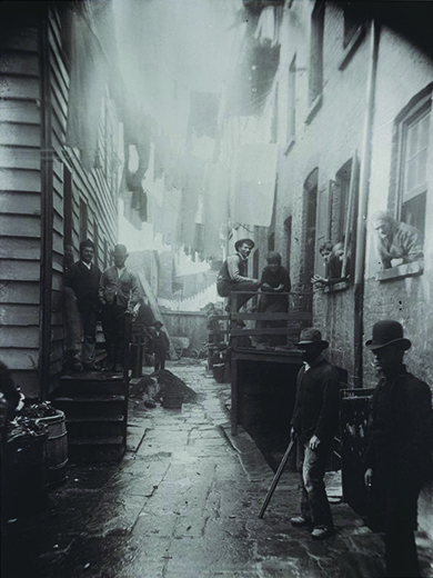 A photograph shows an alley between two tenements. Men, women, and children stand on either side of the alley, in the stoops, and in the windows.