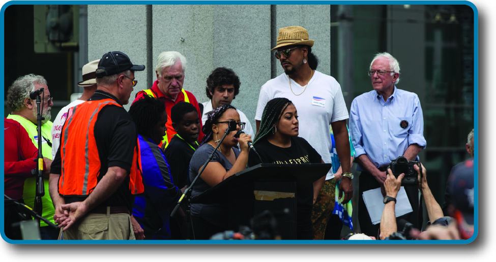 An image of Bernie Sanders on a stage. Bernie is standing to the right and several people are standing in front of a podium.