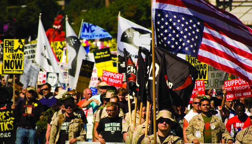 An image of a group of people, several of whom are holding flags and signs. One of the signs reads “End the war now”, and another reads “Support the troops, end the war”.