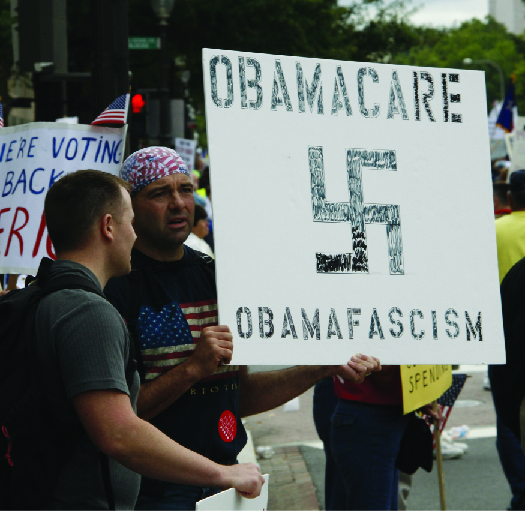 An image of a person holding a sign that reads “Obamacare obamafascism” and has the symbol of a swastika.