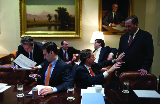 An image of seven people seated around a room. Some are talking to each other. Some are examining papers.