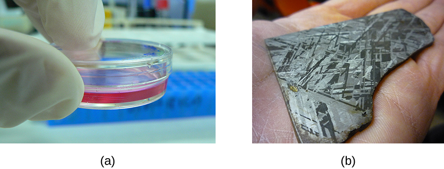 Figure a shows the tips of a person’s gloved hands holding a clear plate with a lid. The plate contains a reddish material in the bottom of the plate. Figure b shows a piece of metal in a person’s hands. The material is dark with some shiny regions.